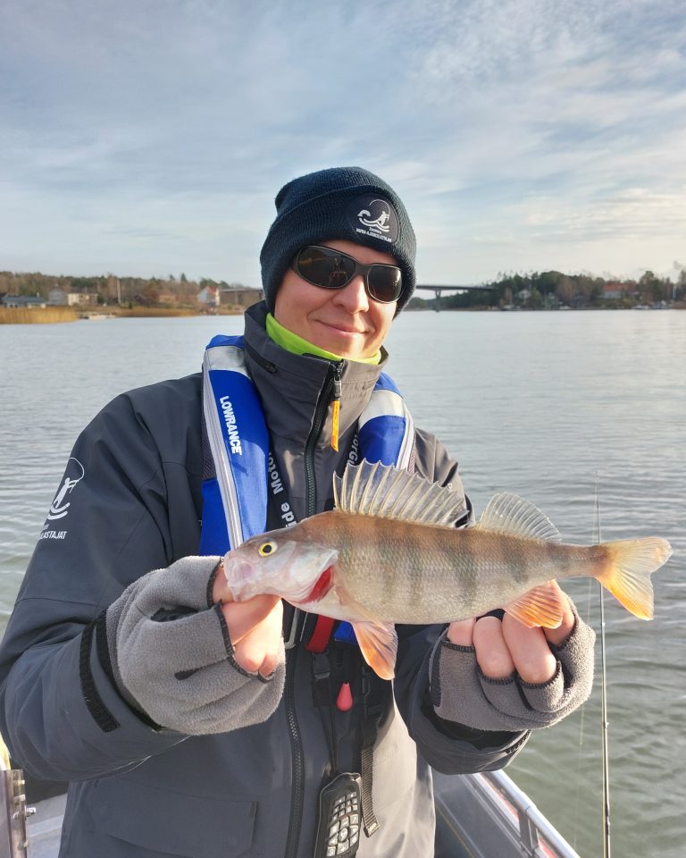 Hymyilevä pipopäinen kalastaja esittelee kalastamaansa ahventa.