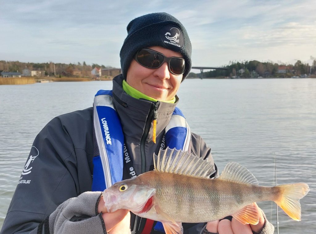 Hymyilevä pipopäinen kalastaja esittelee kalastamaansa ahventa.